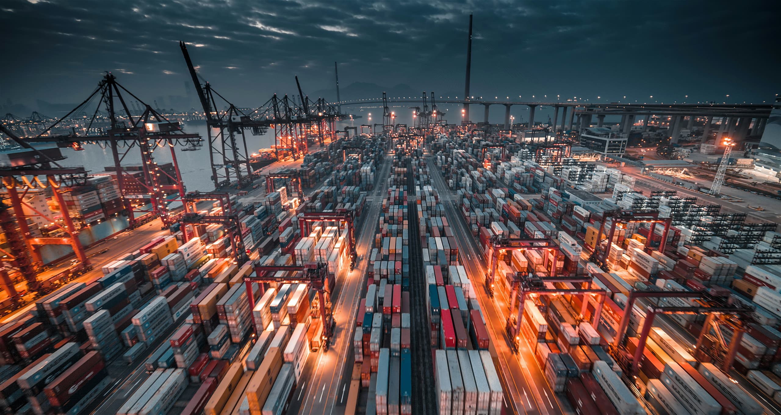 Aerial view of a container terminal at dusk, with stacked shipping containers, gantry cranes and a bridge beyond